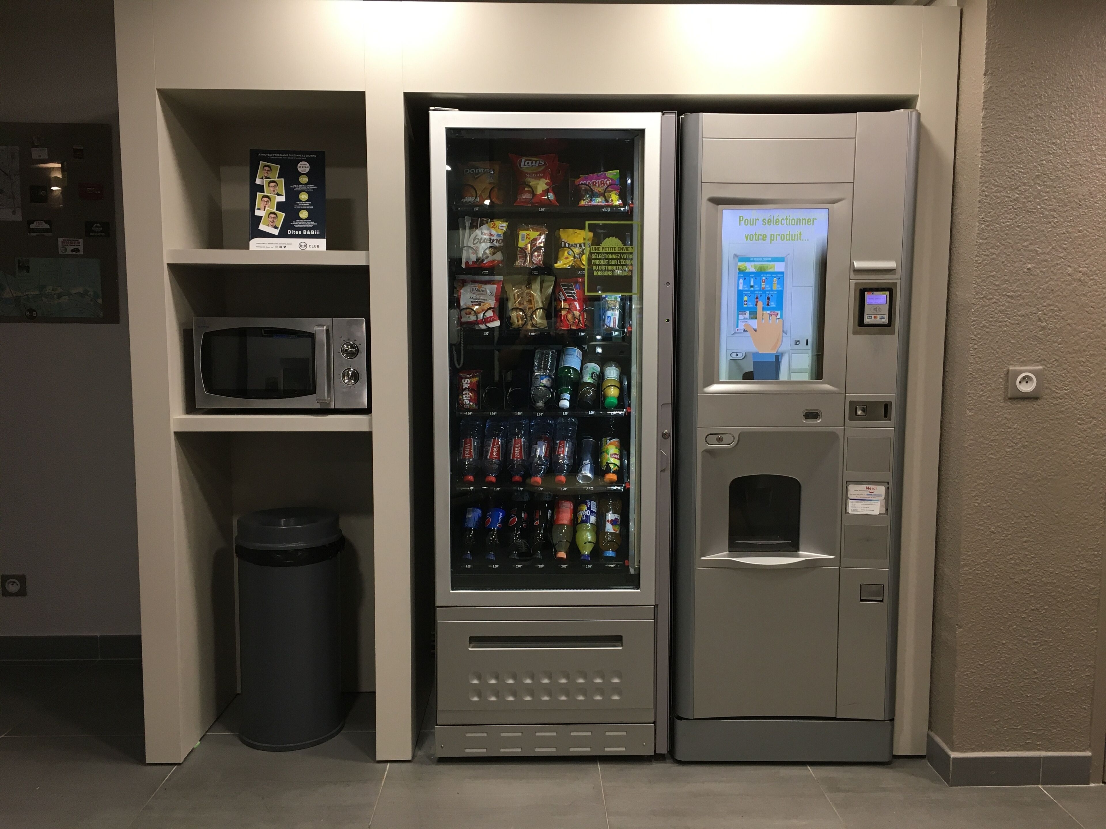 A vending machine with a variety of snacks and drinks, a trash bin, and a microwave above it.