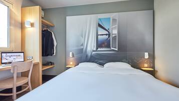 Une chambre d’hôtel avec un lit, un bureau avec un ordinateur, une chaise, une armoire et une grande fenêtre donnant sur un pont.