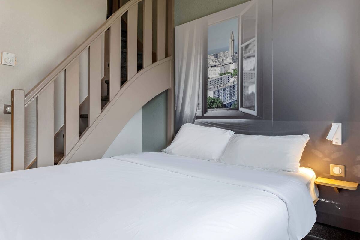Een hotelkamer met een bed, uitzicht op een stadsbeeld door een open raam, een trap en een nachtkastje met een lamp.