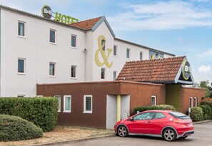 Un hôtel dont le logo, constitué d’un « & » bien visible, est mis en valeur, avec une voiture rouge garée devant et une haie bien entretenue.