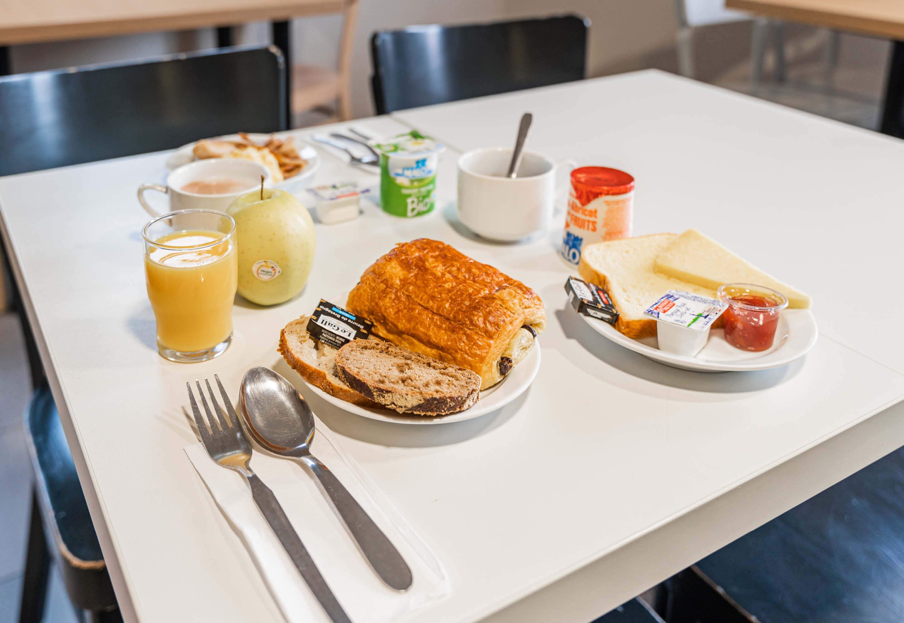 A breakfast setting with a croissant, bread, cheese, jam, butter, and a cup of coffee on a white table.