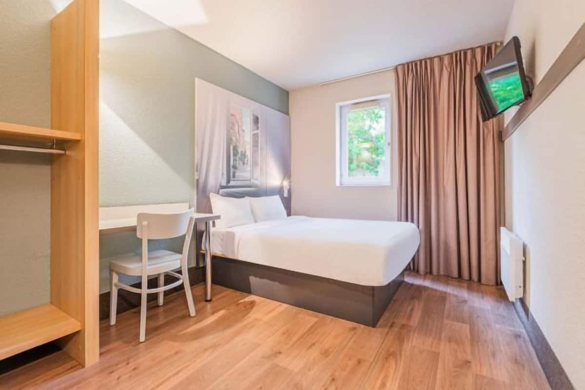 Een kleine, moderne kamer met een bed, een bureau, een stoel en een aan de muur bevestigde televisie.