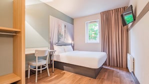 Een slaapkamer met een bed, een bureau, een stoel, een aan de muur bevestigde televisie en een raam met gordijnen.