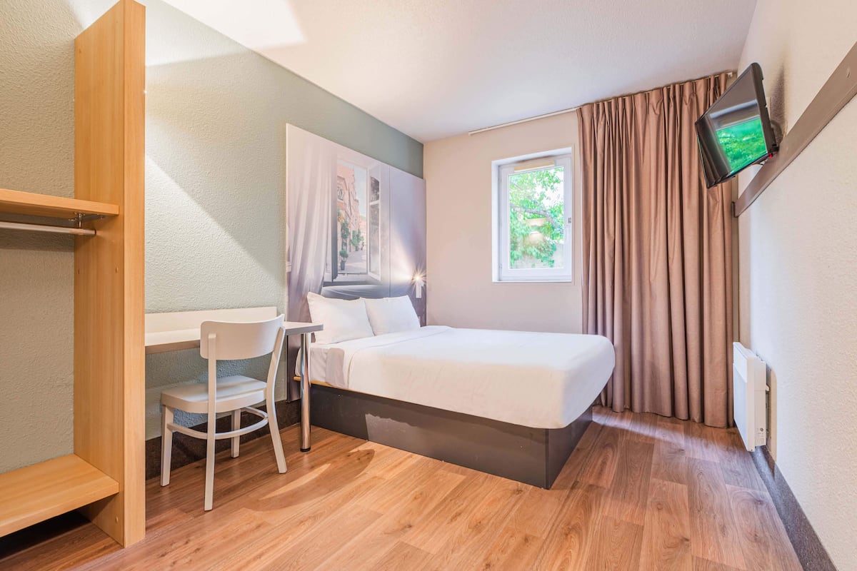 Een slaapkamer met een bed, een bureau, een stoel, een aan de muur bevestigde televisie en een raam met gordijnen.