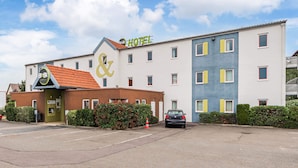 Een meerlaags hotel met een opvallend '&' logo op de gevel, een parkeerplaats en een rode auto ervoor geparkeerd.
