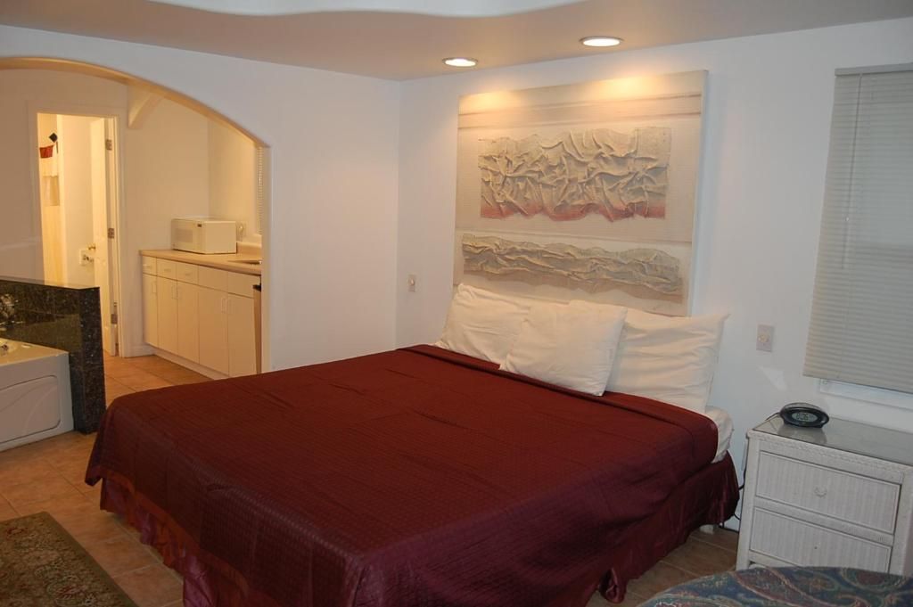 A bedroom with a bed, a nightstand, a wall-mounted artwork, and a bathroom visible through an archway.