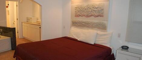 A bedroom with a bed, a nightstand, a wall-mounted artwork, and a bathroom visible through an archway.
