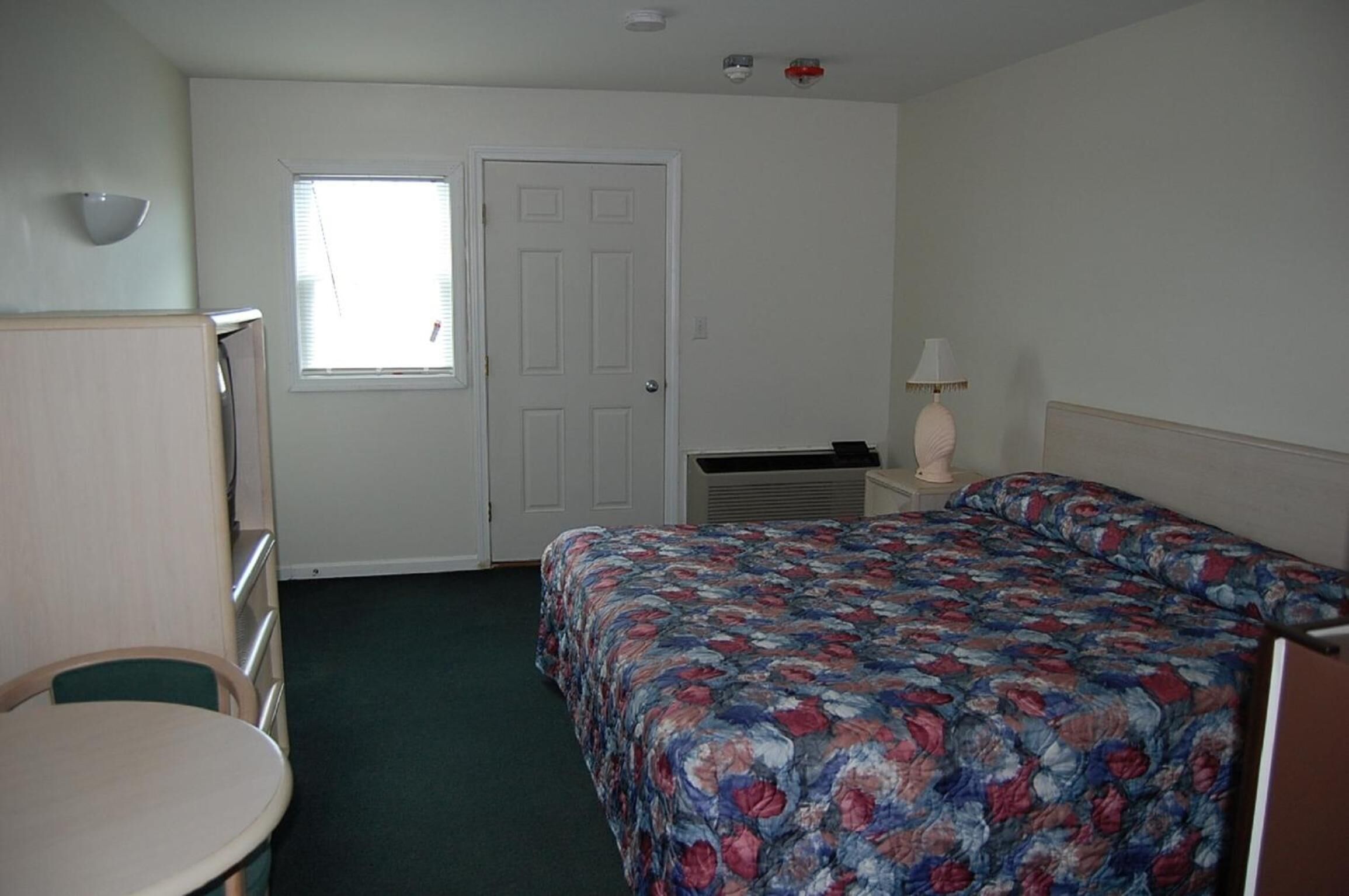 A small, simple room with a bed, a desk, and a chair.