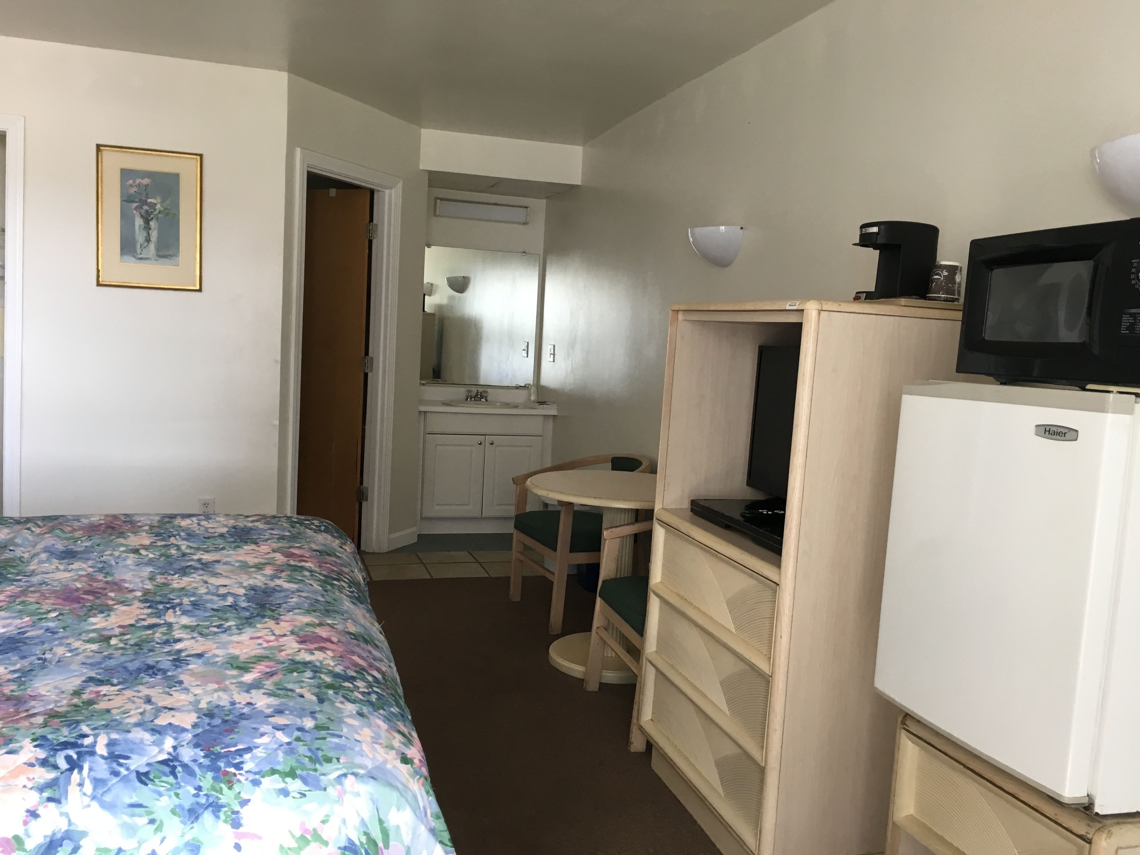 A hotel room with a bed, a small desk, a refrigerator, a microwave, and a bathroom with a sink and mirror.
