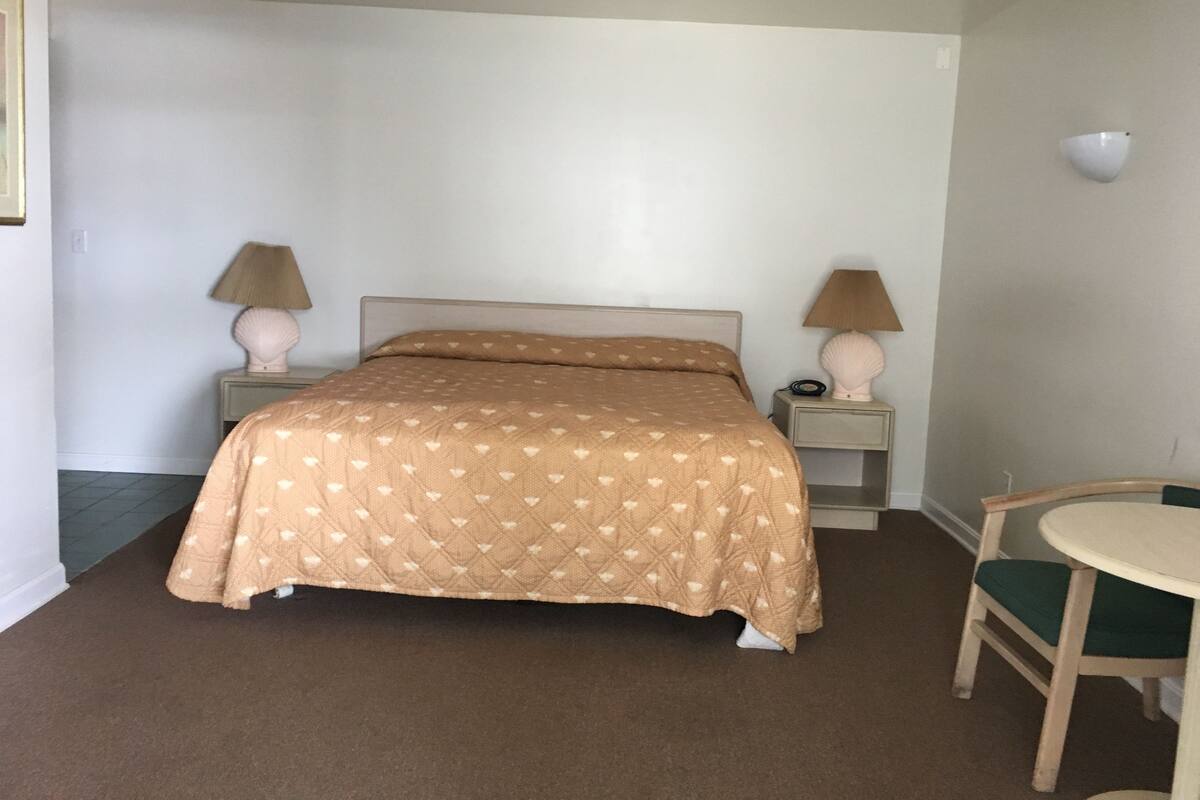 Un dormitorio con una cama, dos mesitas de noche con lámparas, una silla y una pequeña mesa.