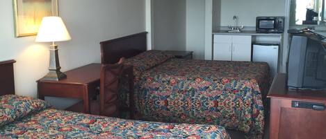 A hotel room with two beds, a desk, a chair, a lamp, a television, and a kitchenette.
