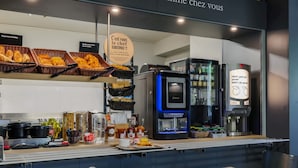 Un bufé de desayuno con panes y pasteles variados, una máquina de café y un cartel dando la bienvenida a los clientes.