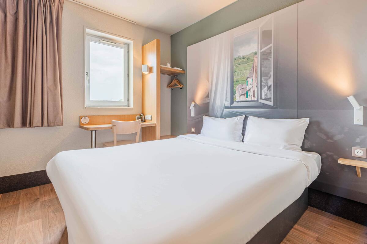 A hotel room with a large bed, a desk, and a chair. There is a window with a view of buildings and a painting on the wall.