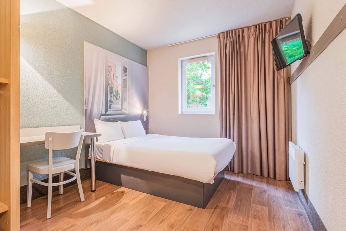 A bedroom with a bed, a desk, a chair, a window with curtains, and a TV mounted on the wall.