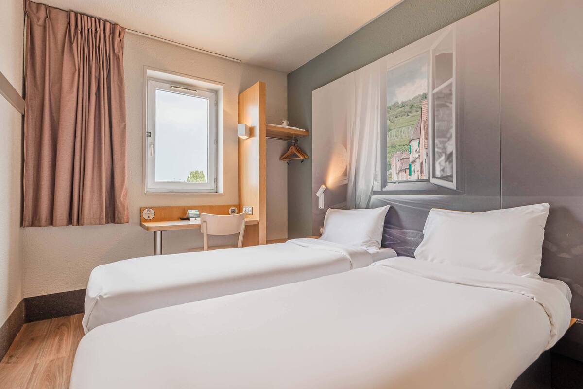 A hotel room with two beds, a desk, a chair, and a large window with curtains.