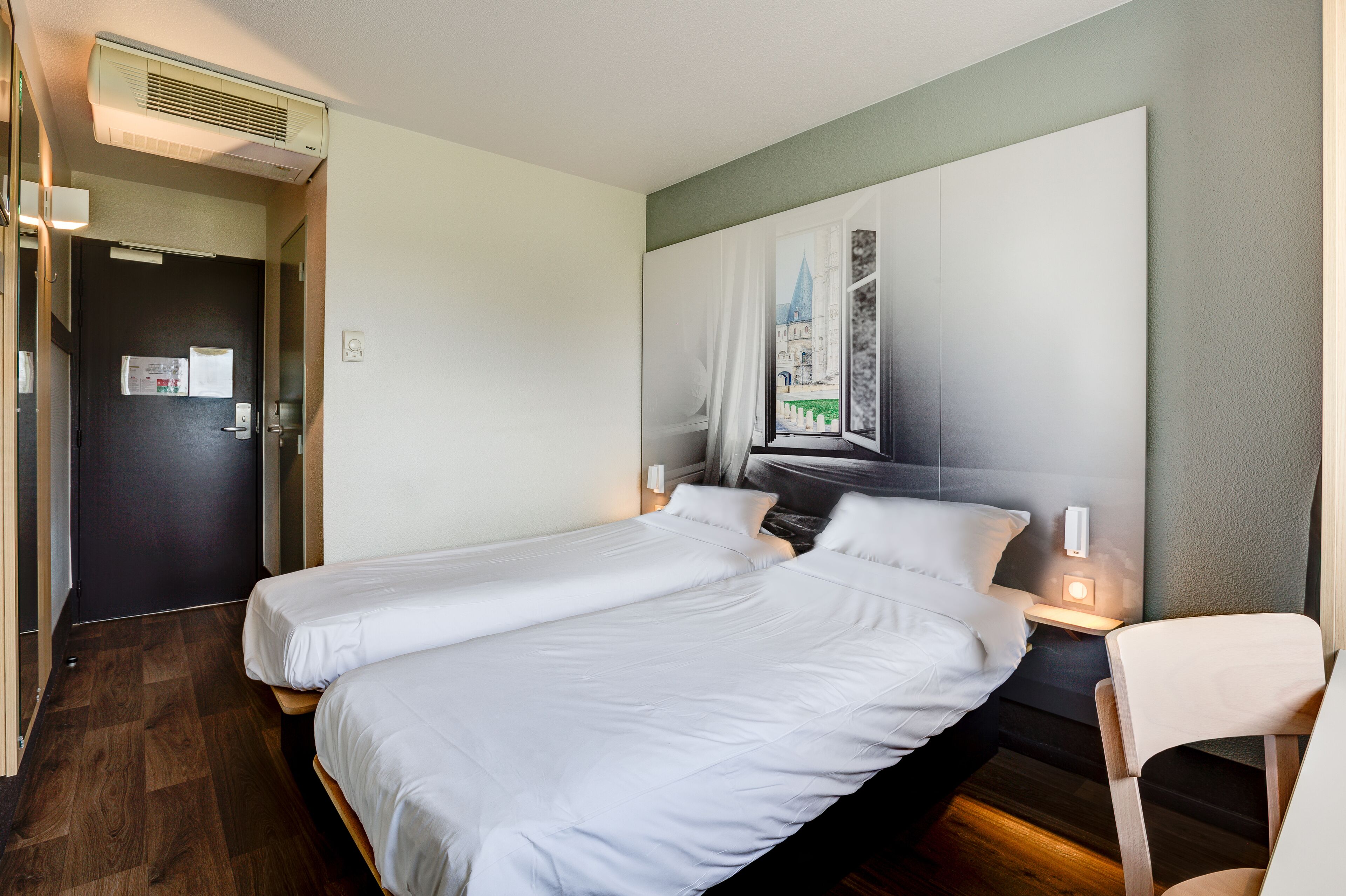 Een hotelkamer met twee enkele bedden, een donkere deur, een schilderij van een kasteel en een tafeltje met een lamp.