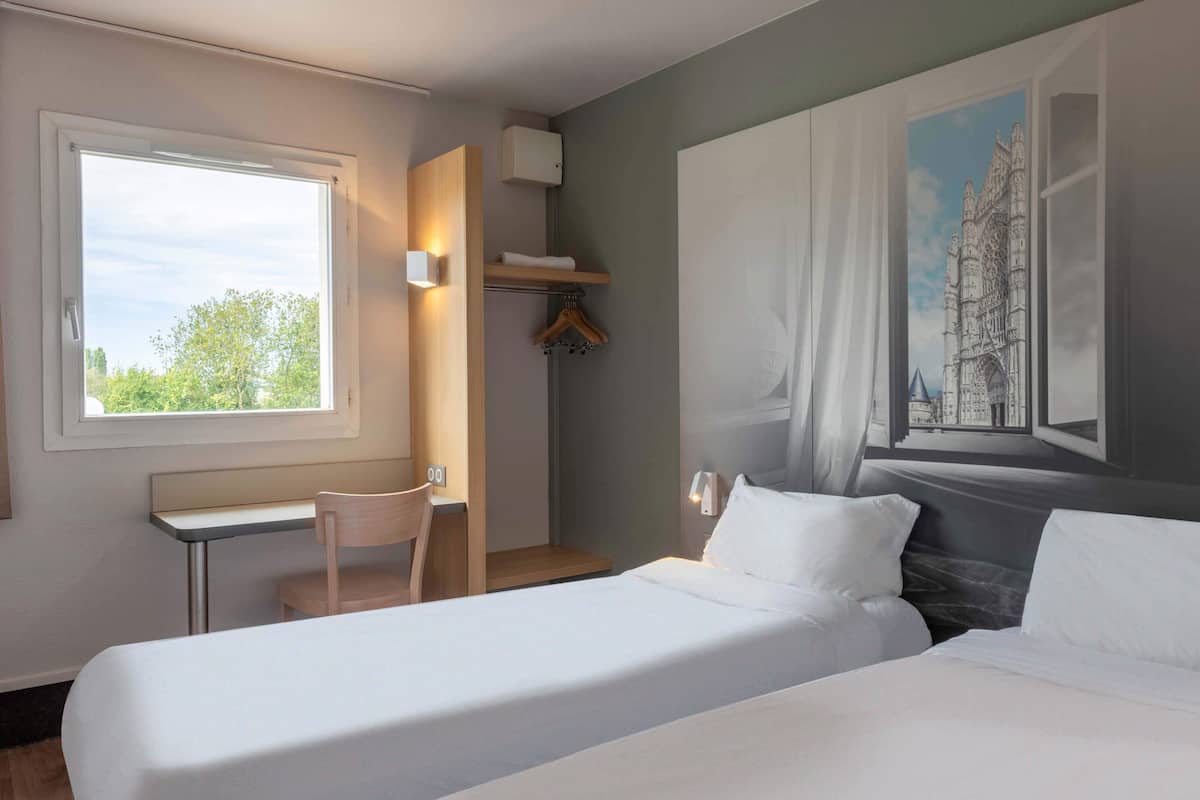 Habitación de hotel con cama, escritorio y silla. Se ve una ventana con vista a árboles y un cuadro de un castillo en la pared.