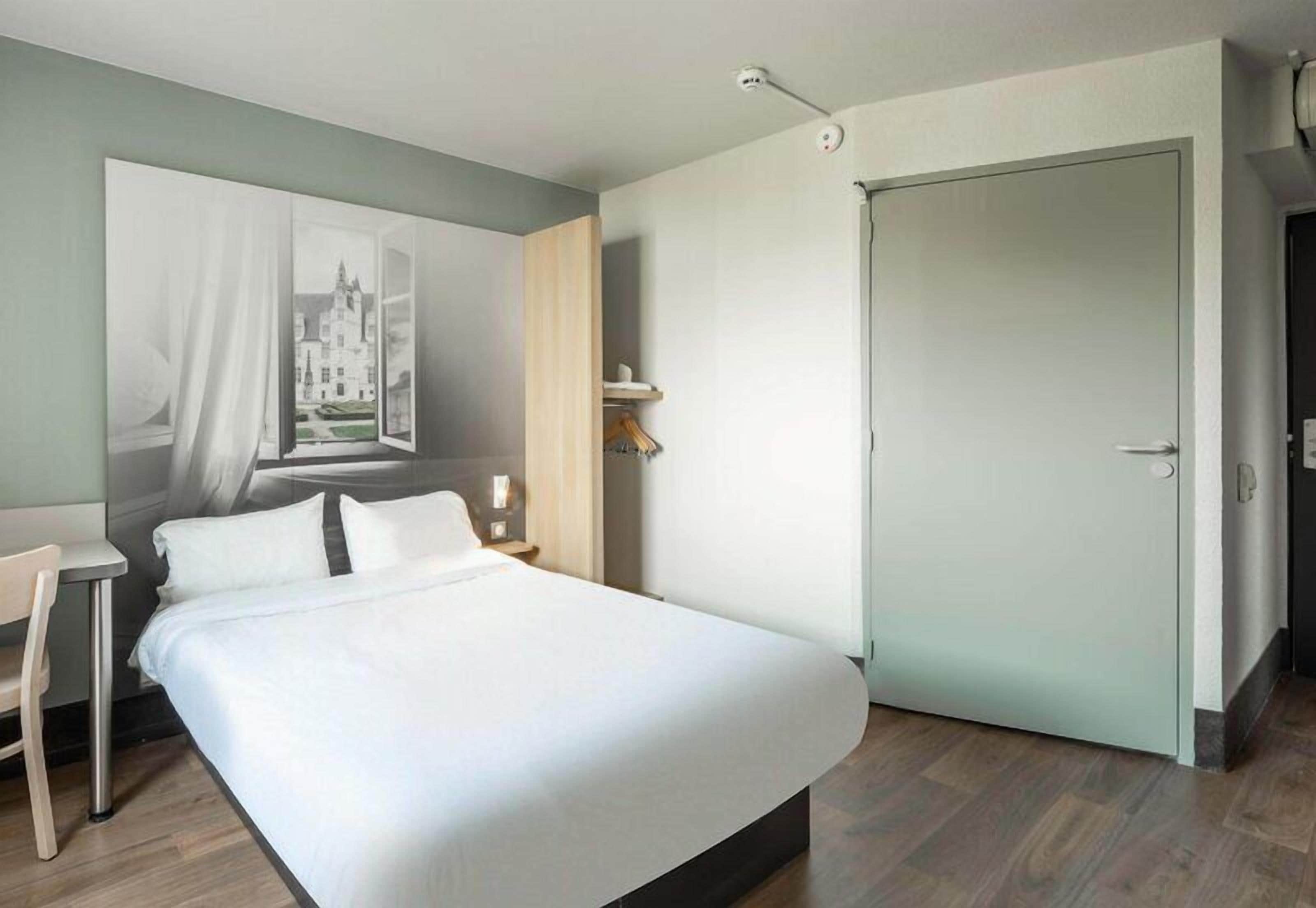 A bedroom with a large bed, a desk, a chair, a door, and a wall-mounted light fixture.