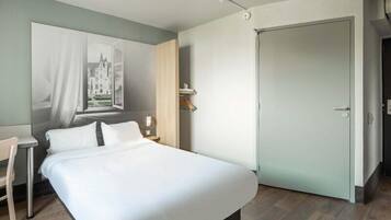 A bedroom with a large bed, a desk, a chair, a door, and a wall-mounted light fixture.
