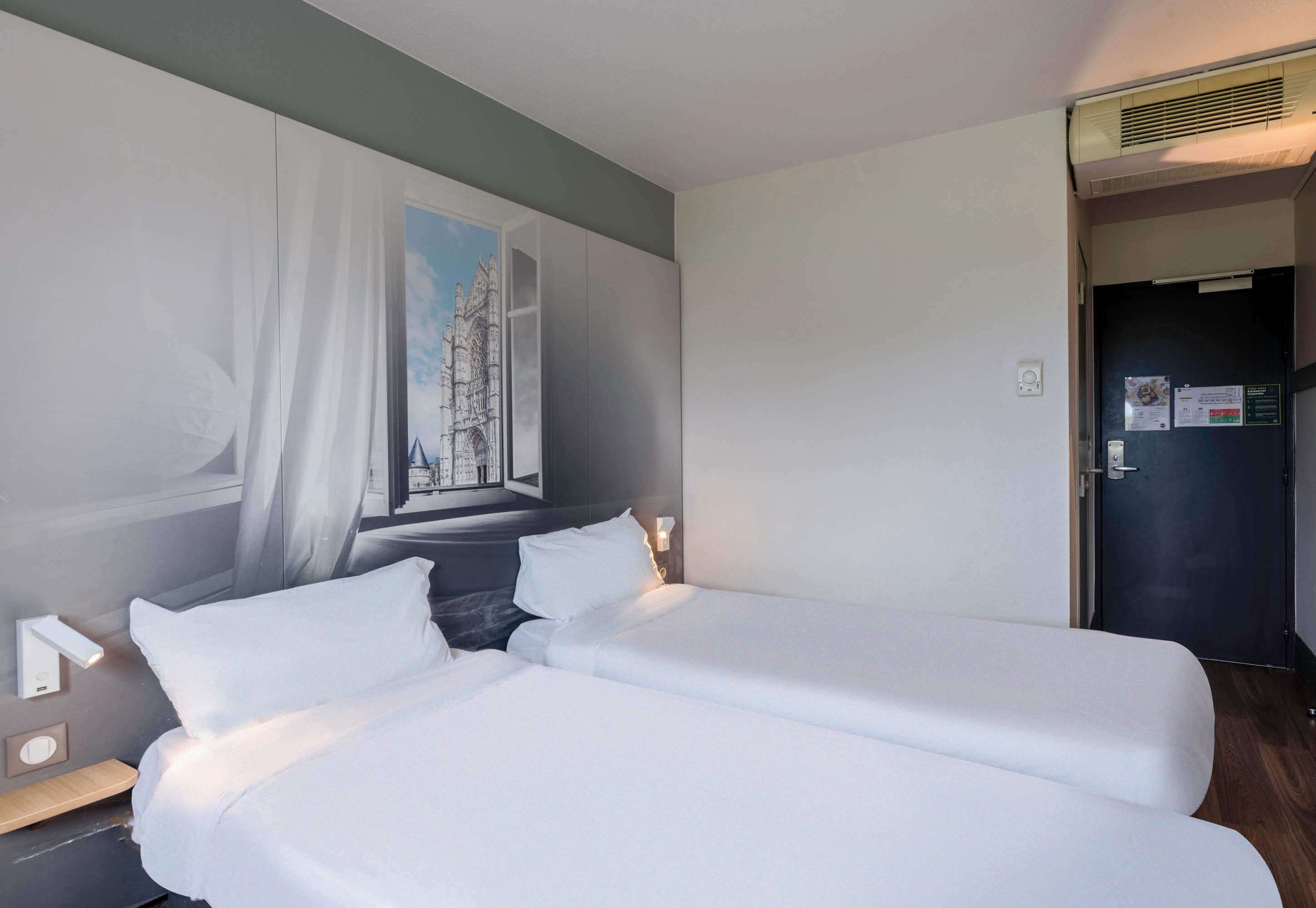 Une chambre d’hôtel avec un grand lit, une fenêtre donnant sur un immeuble et une porte avec une pancarte.