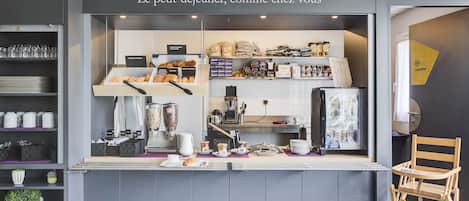 A café with a counter displaying pastries and coffee, a seating area with a chair, and shelves with kitchenware.