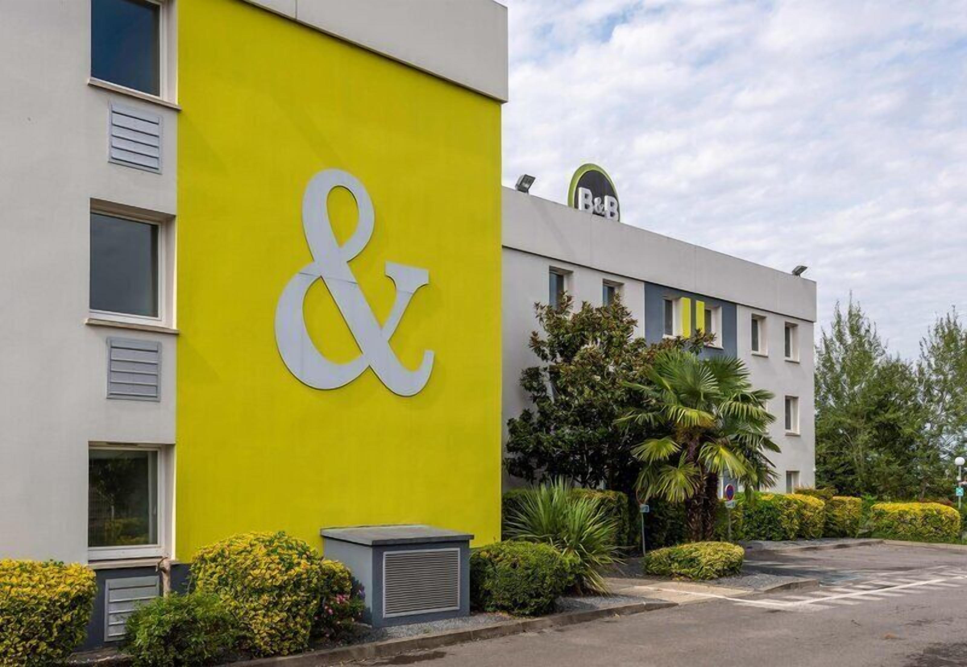 A building with a large '&' sign on a yellow wall, a B&B logo, and a parking lot with greenery.