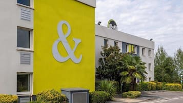 A building with a large '&' sign on a yellow wall, a B&B logo, and a parking lot with greenery.