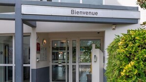 A building entrance labeled 'Bienvenue' with glass doors, a small outdoor seating area, and a security camera.