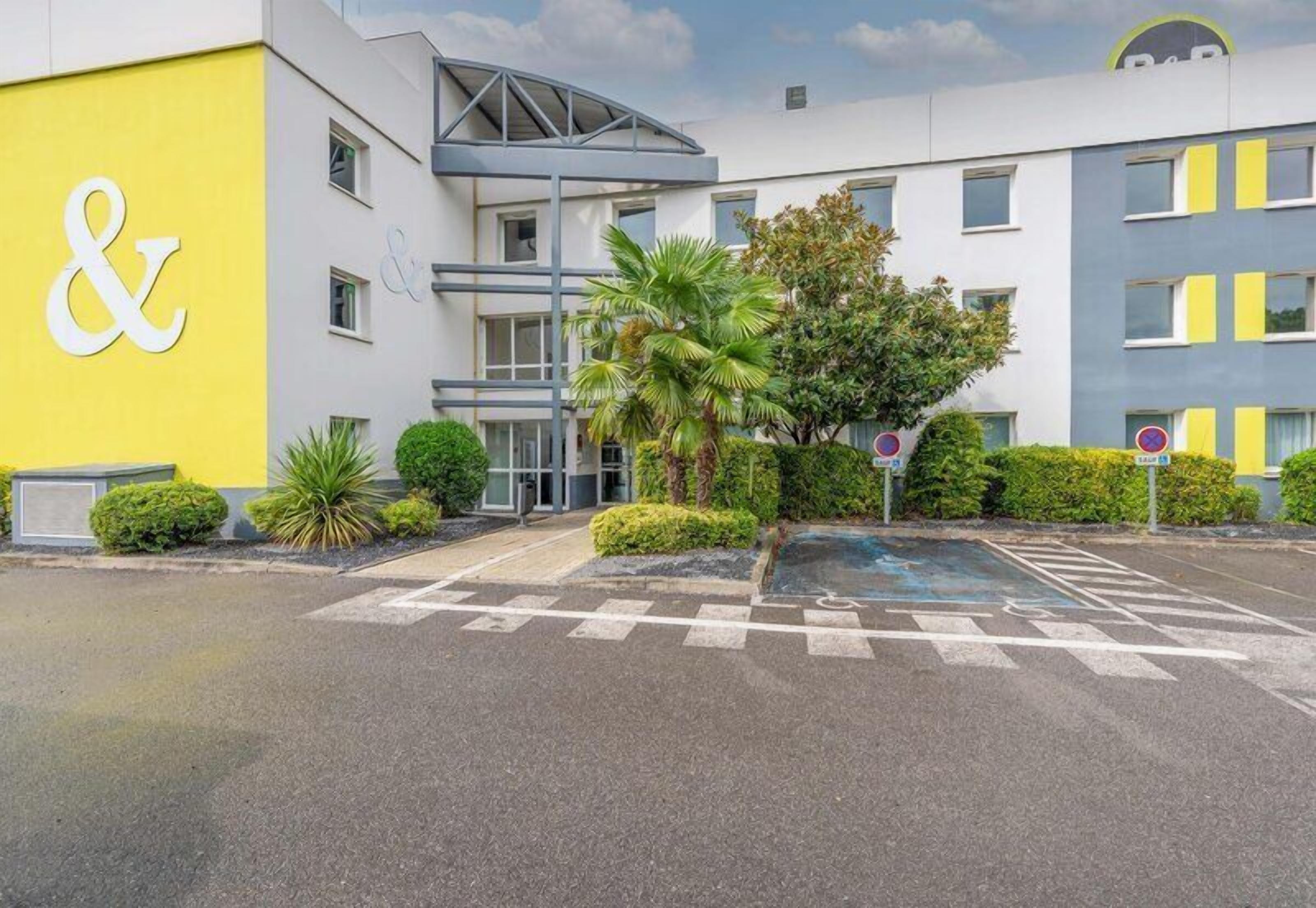 A modern hotel with a distinctive yellow and white facade, a covered entrance, and a parking area.