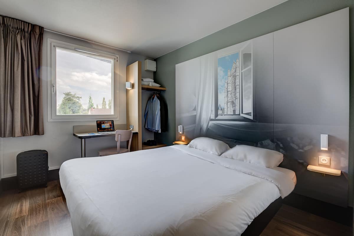 Una habitación de hotel moderna con una cama grande, un escritorio con silla, un armario y un amplio ventanal con cortinas.