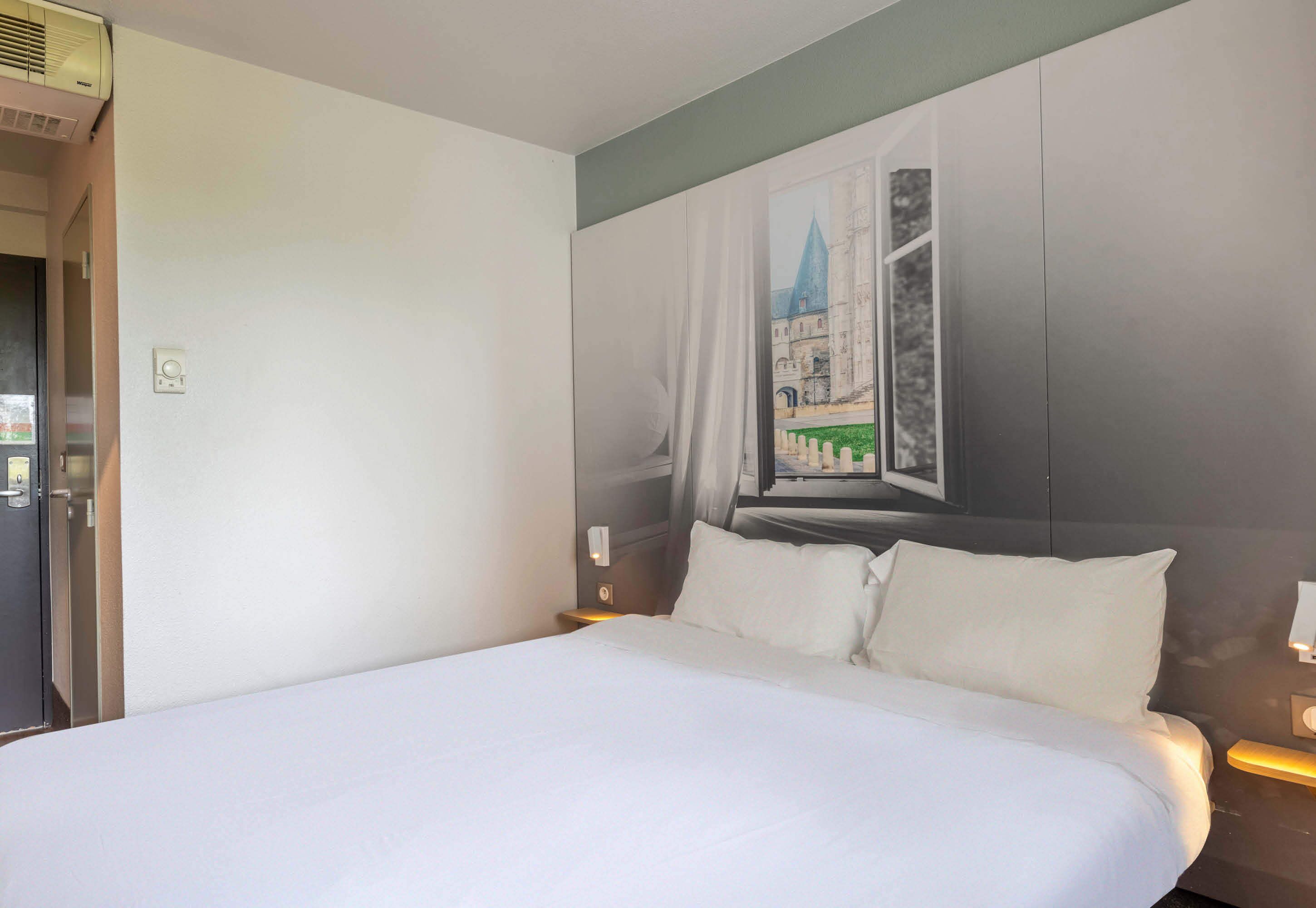 Une chambre d’hôtel avec un grand lit, un tableau représentant un bâtiment accroché au mur et une porte sur le côté gauche.