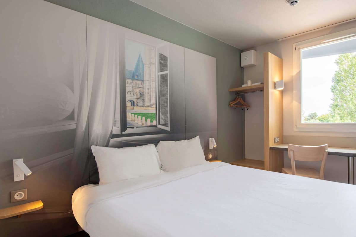 Habitación de hotel con una cama grande, una ventana con vista a un edificio, una silla y un perchero.