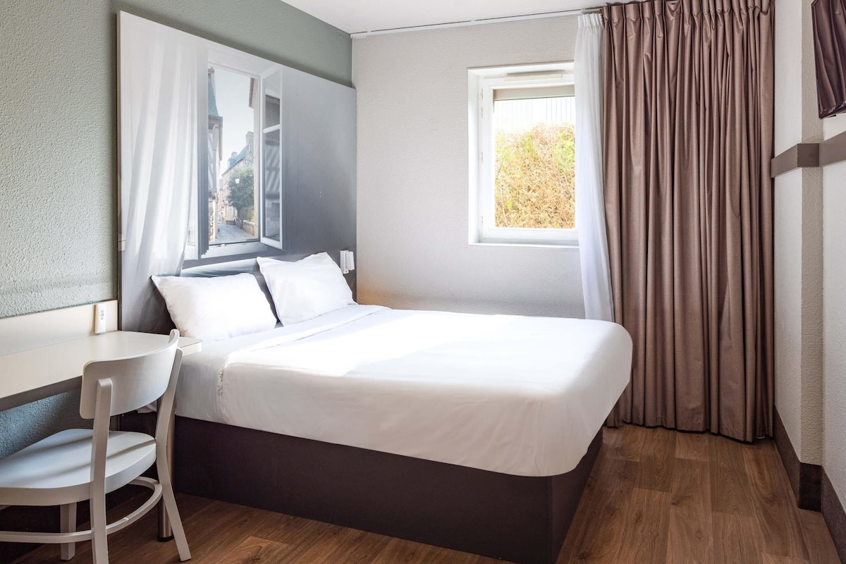 Une chambre d’hôtel avec un lit, un bureau, une chaise, une fenêtre et des rideaux.