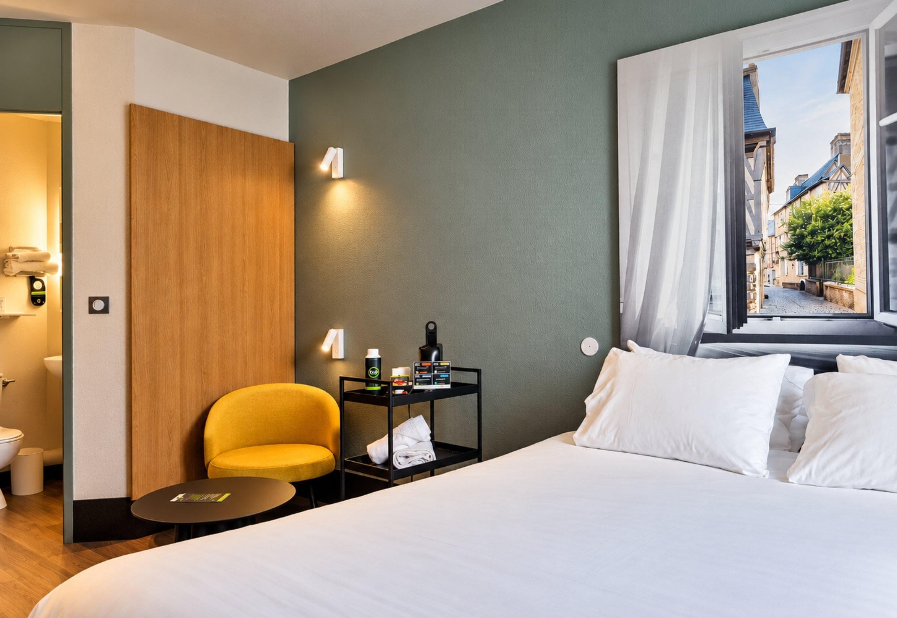 A hotel room with a bed, a small table, a chair, a nightstand with toiletries, and a window with a view of buildings.
