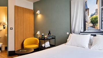 Hotelkamer met een bed, een klein tafeltje, een stoel, een nachtkastje met toiletartikelen en een raam met uitzicht op gebouwen.