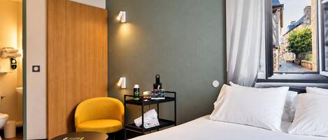 A hotel room with a bed, a small table, a chair, a nightstand with toiletries, and a window with a view of buildings.