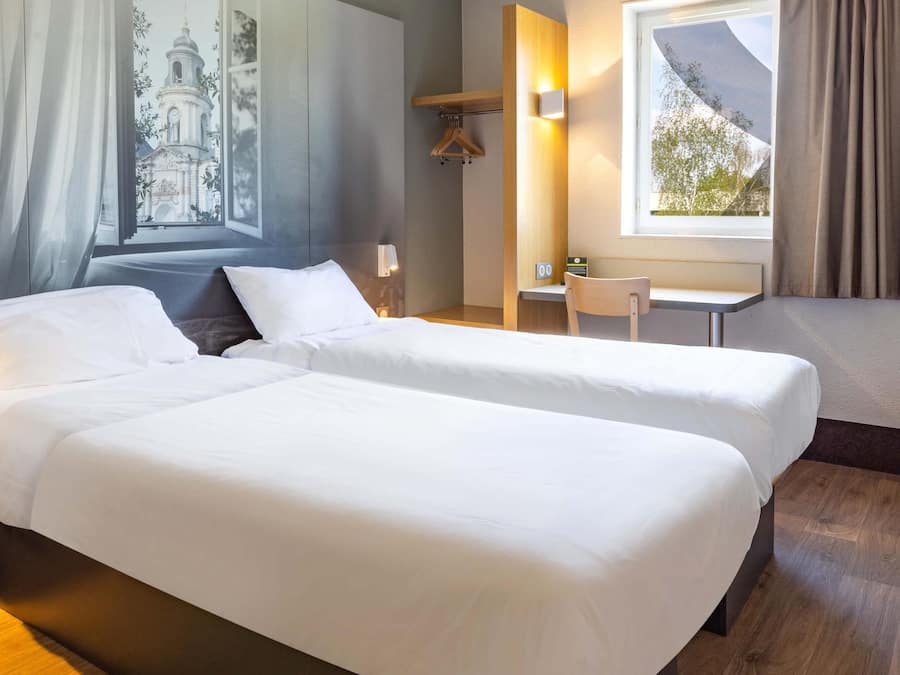 Habitación de hotel con dos camas, un escritorio, una silla, una ventana con vista a una iglesia y una lámpara fijada a la pared.