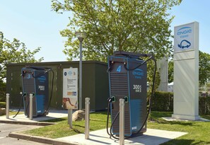 Two electric vehicle charging stations with a sign indicating 300 kW2 capacity, a lamp post, and a tree in the background.