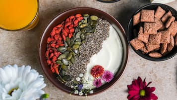 A bowl of yogurt topped with chia seeds, pumpkin seeds, and goji berries, surrounded by flowers and a glass of orange juice.