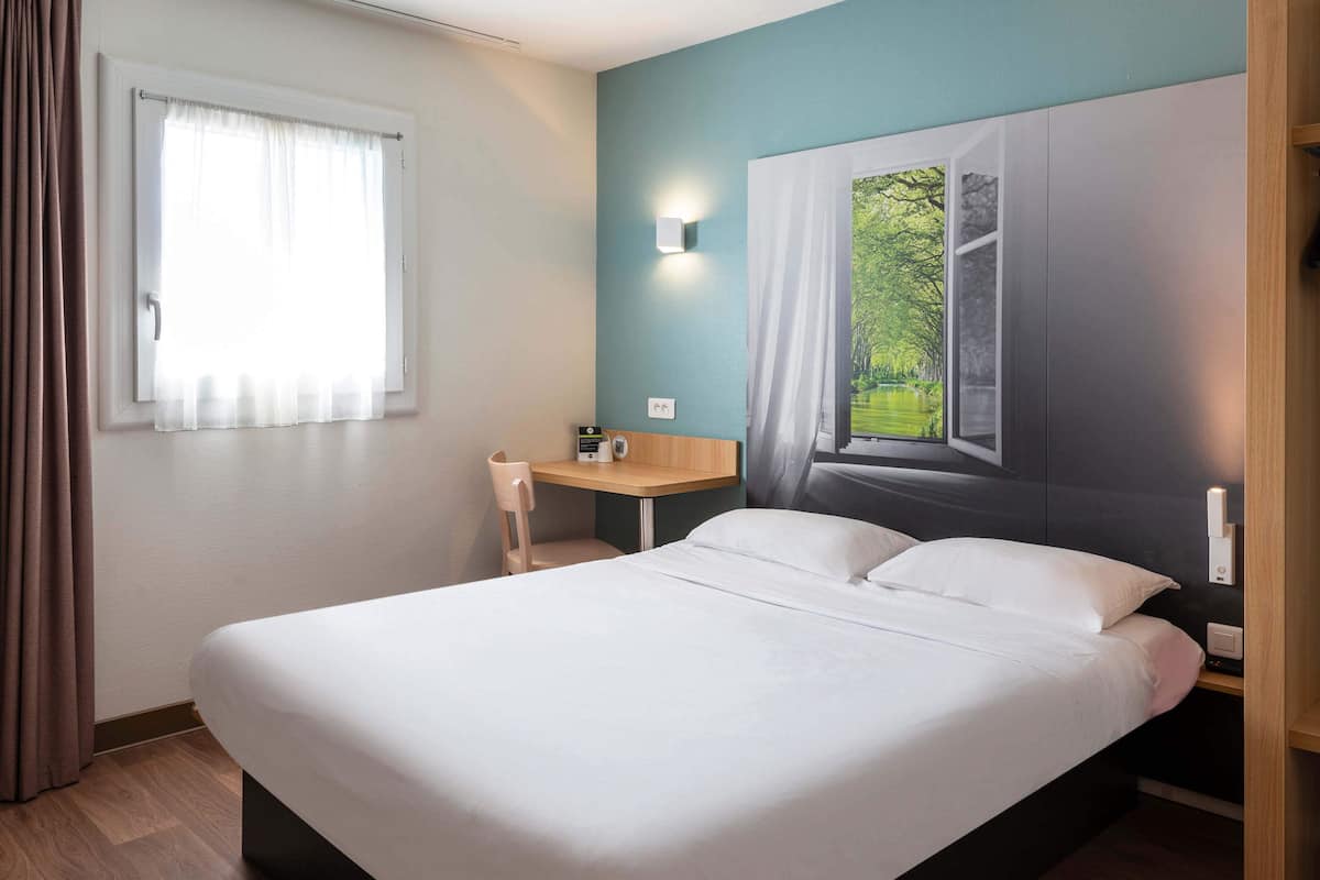 Habitación de hotel con cama, escritorio, silla, ventana con cortinas y una lámpara de pared.