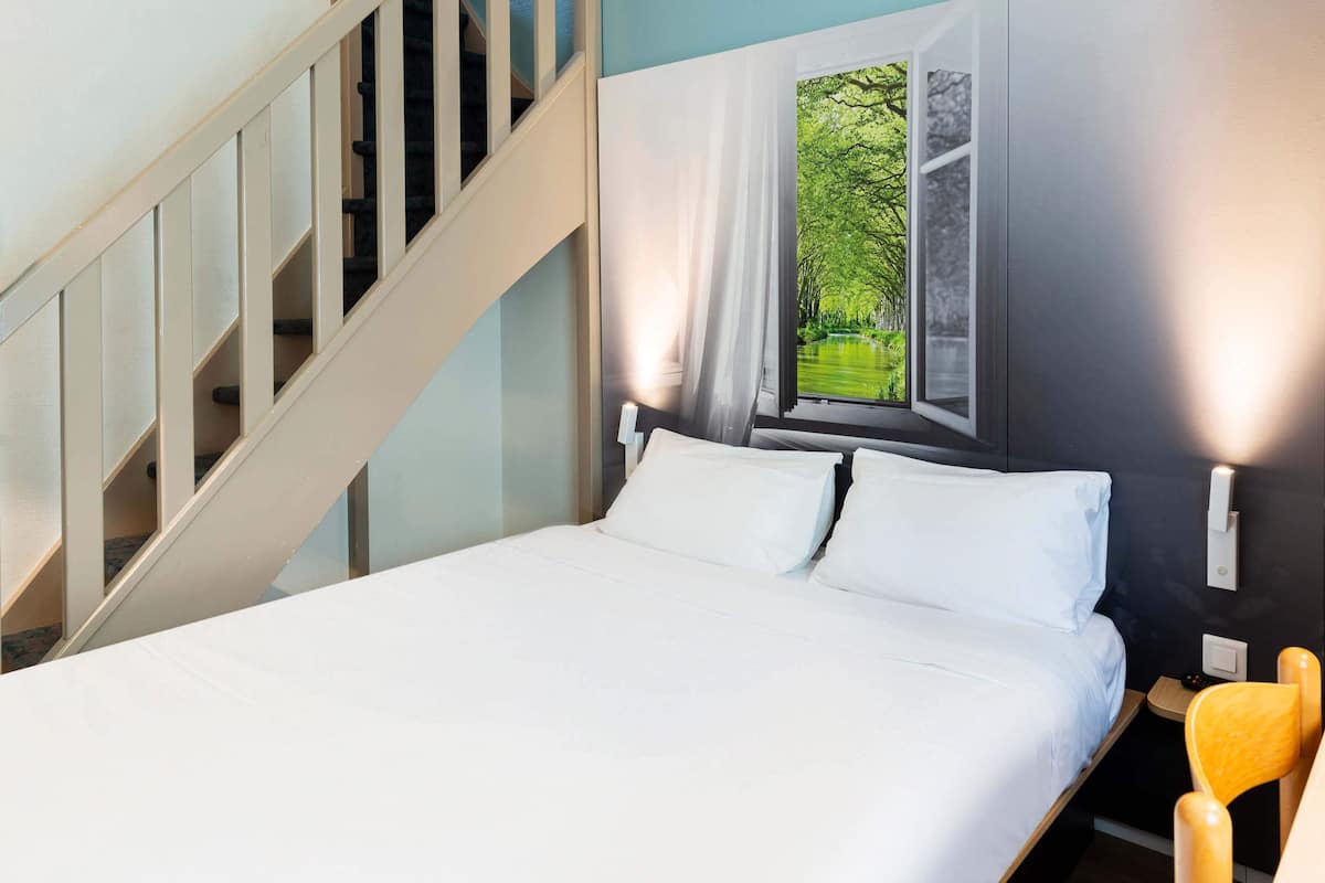 Un dormitorio con cama, ventana con vista a follaje, escalera y mesita de noche con lámpara.