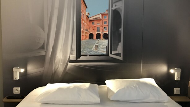 Hotelkamer met een bed, witte kussens, een nachtkastje met een afstandsbediening en uitzicht op een rood gebouw door een open raam.