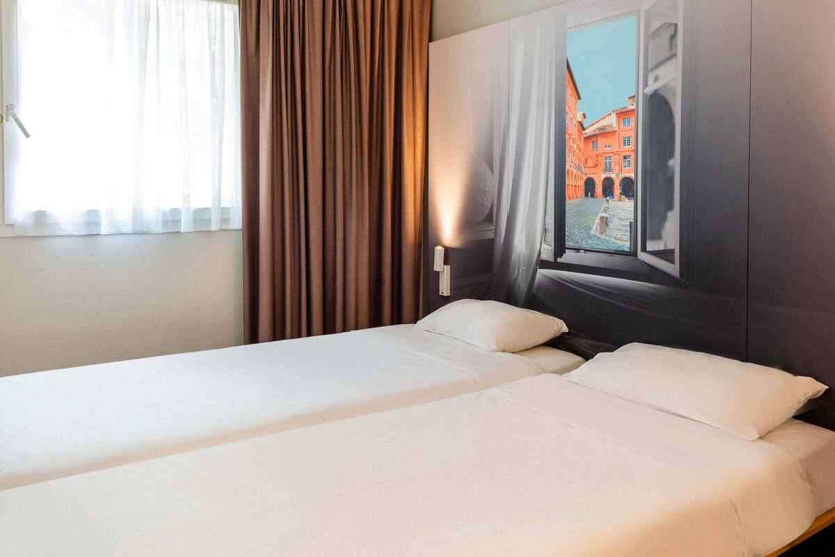 Habitación de hotel con dos camas, una ventana con cortinas y un mural en la pared de un edificio.