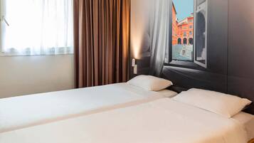 A hotel room with two beds, a window with curtains, and a wall mural of a building.
