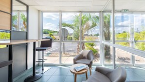 Un área de espera moderna con un escritorio, dos sillas y ventanas amplias que dan a vistas de palmeras y un estacionamiento.