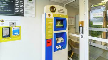 Ein Empfangsbereich eines B&B Hotels mit einem Self-Service-Terminal zur Zimmerbuchung, einer wandmontierten Infotafel und einer Tür mit Glasfenster.