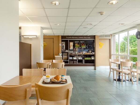 A café with wooden tables and chairs, a breakfast tray with croissant and orange juice, and a view of greenery outside. - B&B HOTEL Creil Chantilly (Creil)