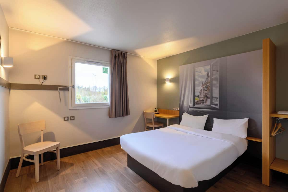 Een hotelkamer met een bed, een bureau, een stoel, een raam met gordijnen en een schilderij aan de muur.