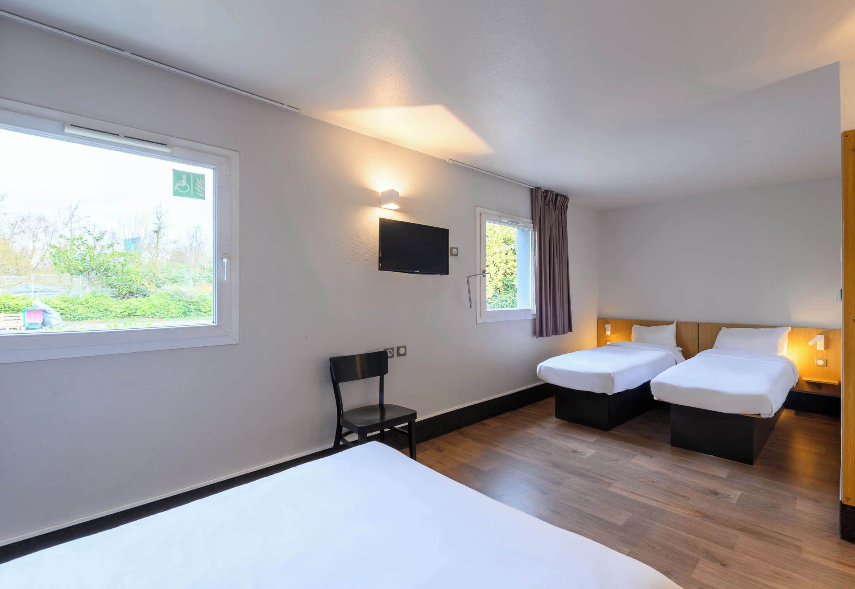 Habitación de hotel con dos camas individuales, un escritorio, una silla, un televisor y una ventana con vistas a zonas verdes.
