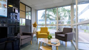Un vestíbulo moderno con un sillón amarillo, dos sillones grises, una mesita con una planta y una máquina expendedora.